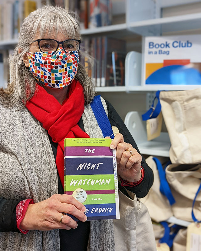 A library staff member stands in front of the Book Club in a Bag shelf and holds up a paperback copy of The Night Watchman