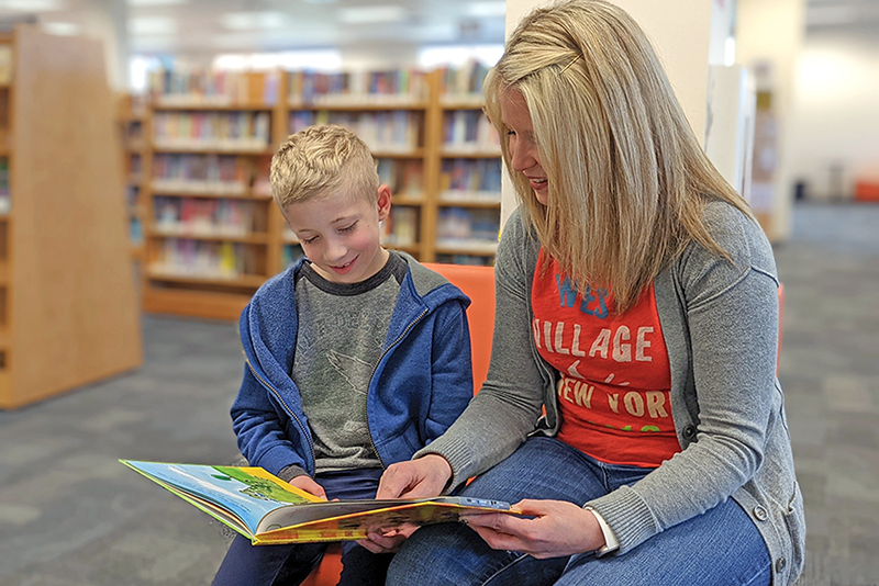 A young boy sits beside his mother in the library smiling at a hardcover book on their laps